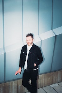 a man in a black jacket and jeans standing in front of a wall